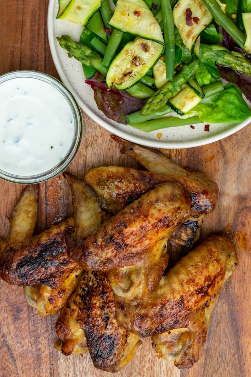 Baked chicken wings on a wooden board with plate of vegetables and sauce top view